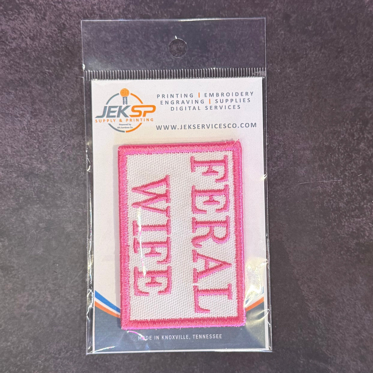 Feral Wife patch in white with pink FERAL WIFE lettering and border, in clear retail packaging