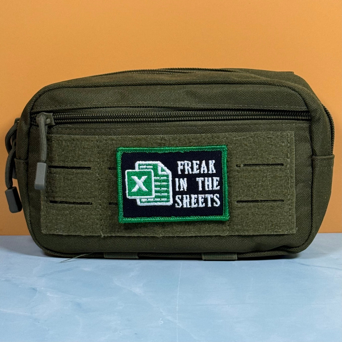 Freak In The Sheets patch in black with a green spreadsheet icon, white lettering and green border, mounted on an olive utility pouch