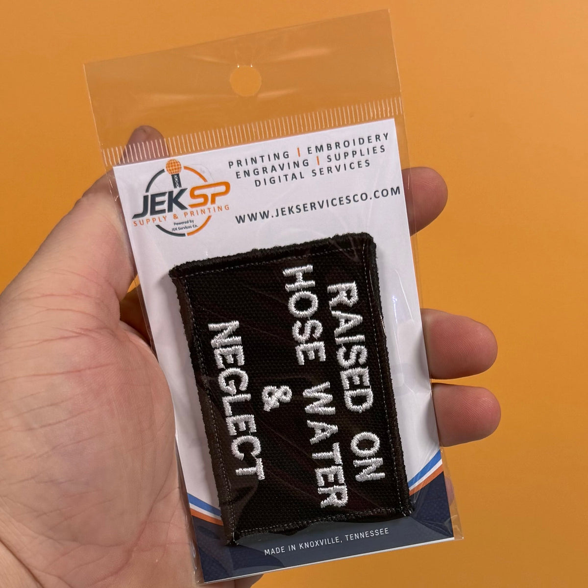 Raised on Hose Water & Neglect patch in black with white lettering, retail package held in a hand