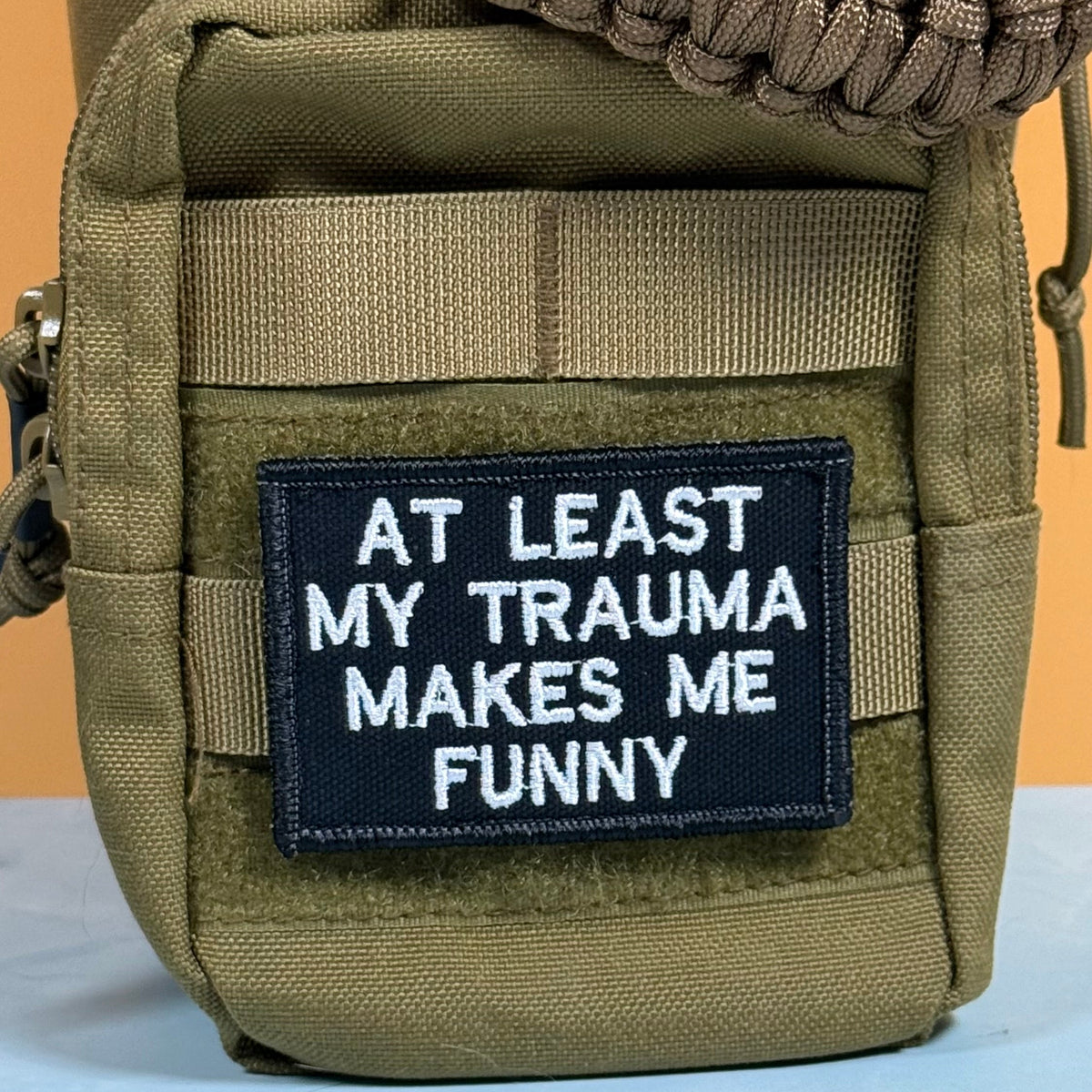 At Least My Trauma Makes Me Funny patch in black with white lettering, mounted on an olive backpack