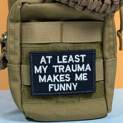At Least My Trauma Makes Me Funny patch in black with white lettering, mounted on an olive backpack