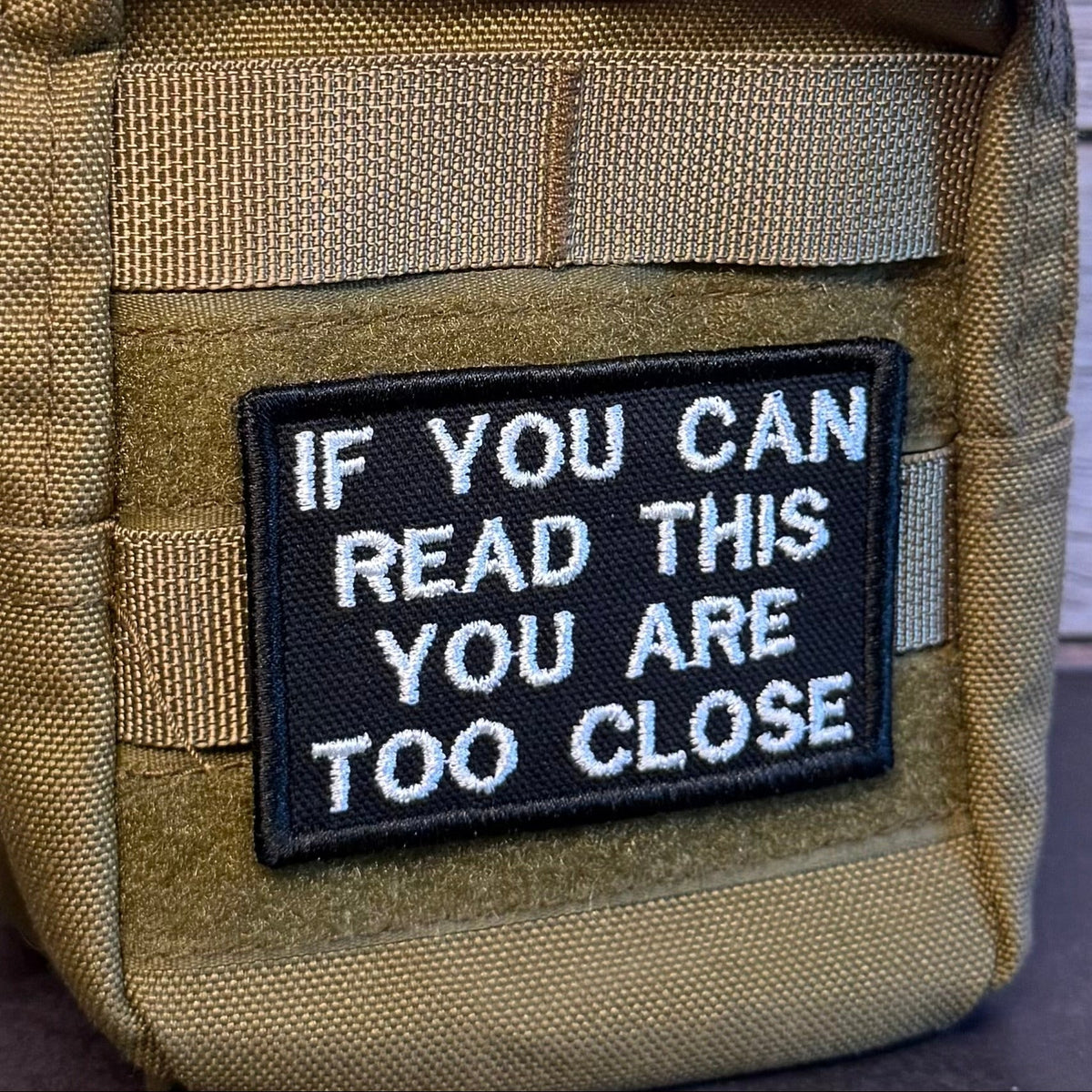 If You Can Read This You Are Too Close patch in black with white lettering, mounted on an olive backpack