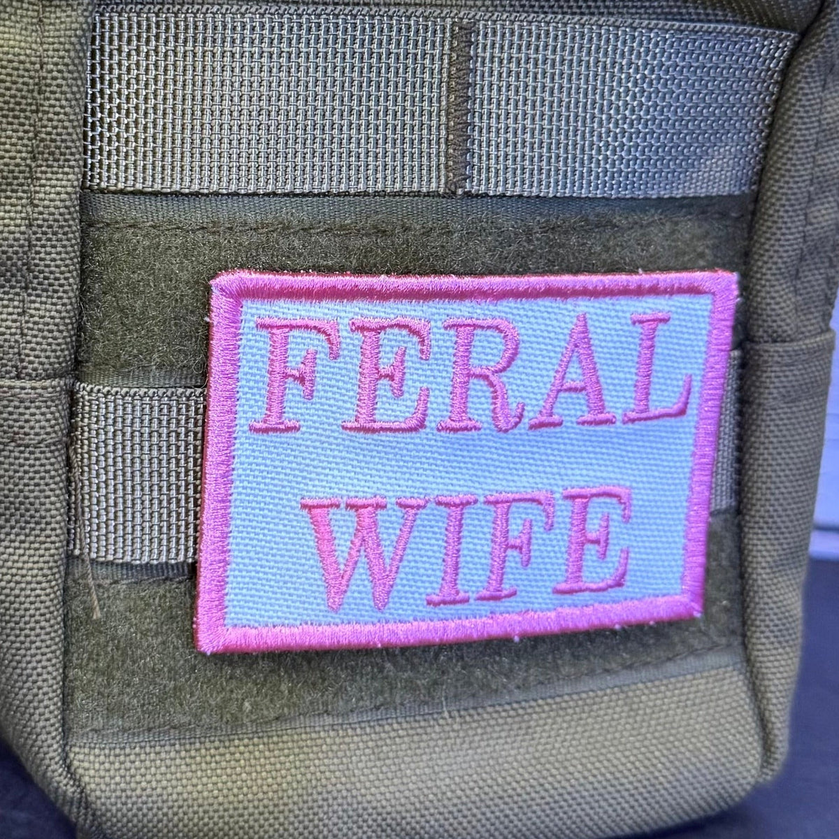 Feral Wife patch in white with pink FERAL WIFE lettering and border, mounted on an olive backpack