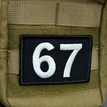 67 (Six Seven) patch in black with white 67 numerals, mounted on an olive backpack