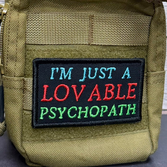 I'm Just A Lovable Psychopath patch in black with blue, red and green lettering, mounted on an olive backpack