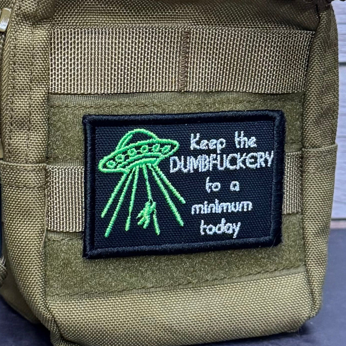 Keep the Dumbfuckery To A Minimum patch in black with a bright green UFO and white lettering, mounted on an olive backpack