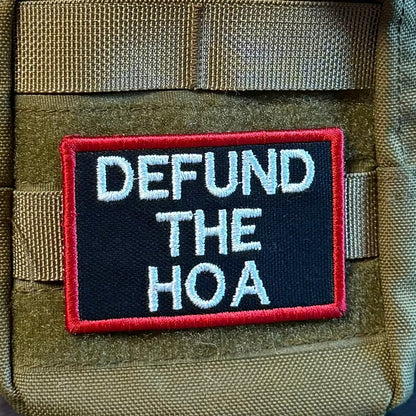 Defund the HOA patch in black with white lettering and a red border, mounted on an olive backpack