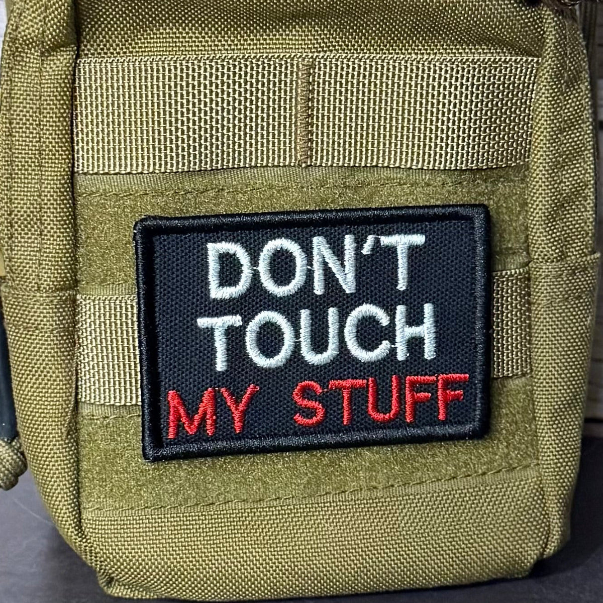 Don't Touch My Stuff patch in black with white and red lettering, mounted on an olive backpack