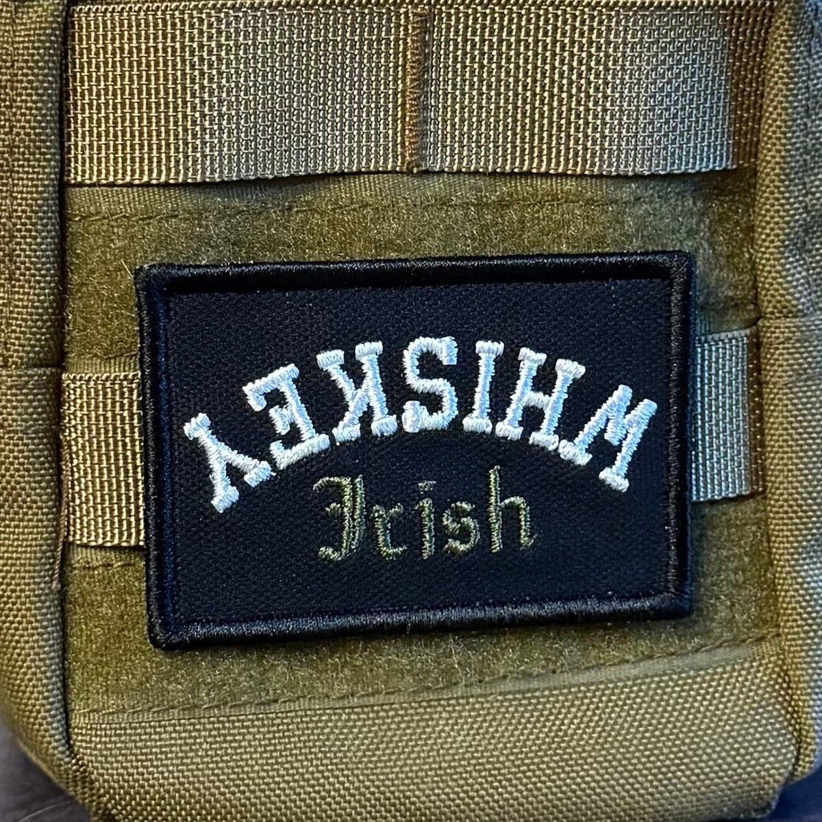 Irish Whiskey Upside Down patch in black with white upside-down WHISKEY lettering and green Irish script, mounted on an olive backpack