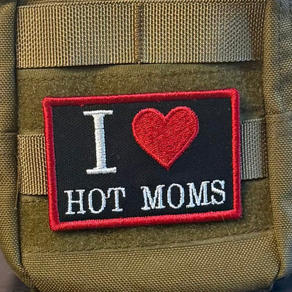 I Love Hot Moms MILFS patch in black with a red heart and border and white lettering, mounted on an olive backpack