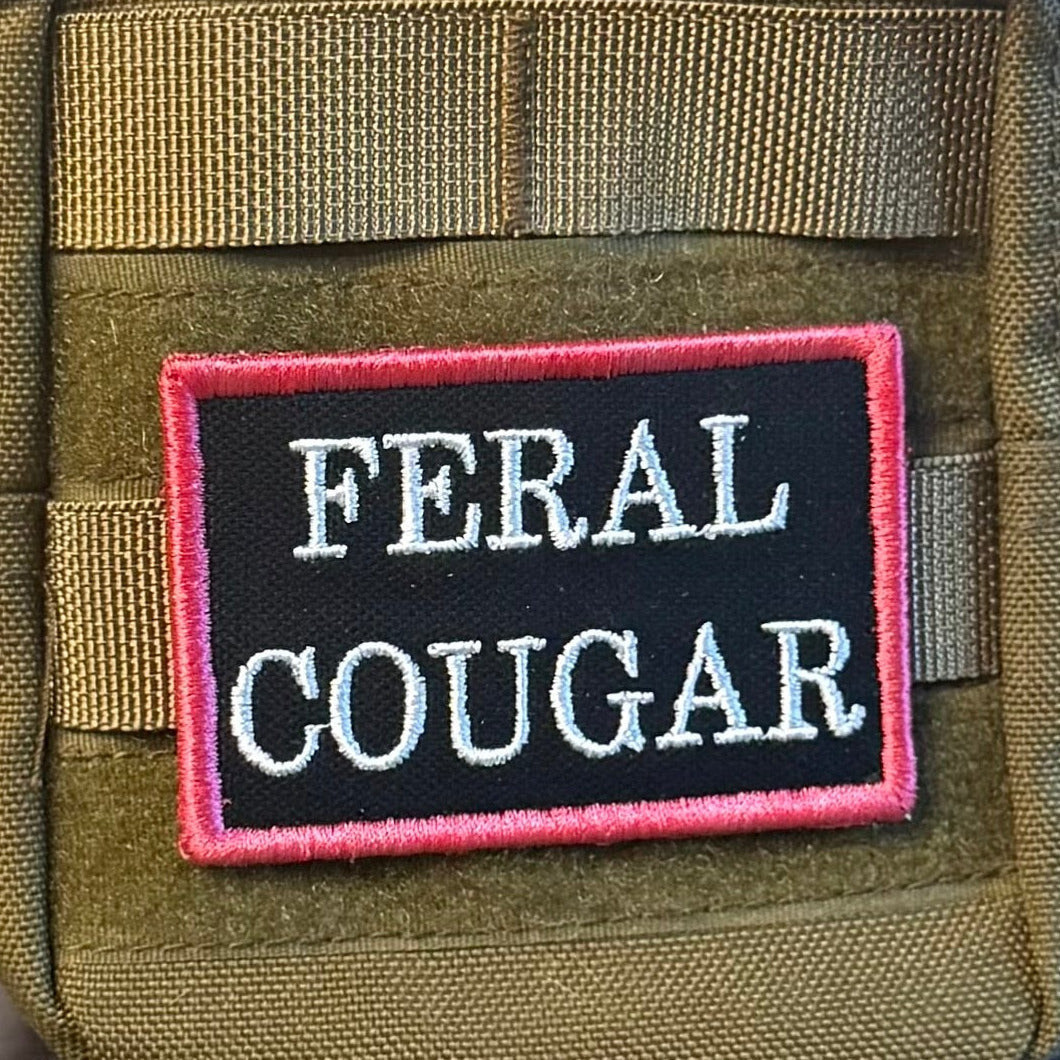 Feral Cougar patch in black with white lettering and pink border, mounted on an olive backpack