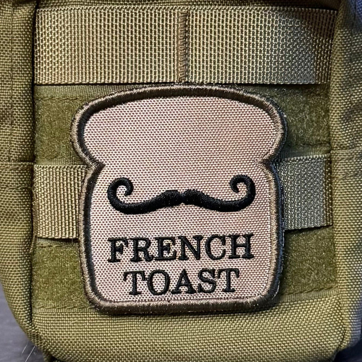 French Toast patch in tan toast-shaped design with a black moustache and lettering, mounted on an olive backpack