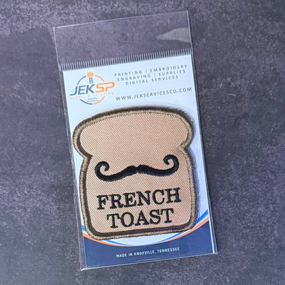 French Toast patch in tan toast-shaped design with a black moustache and lettering, in clear retail packaging
