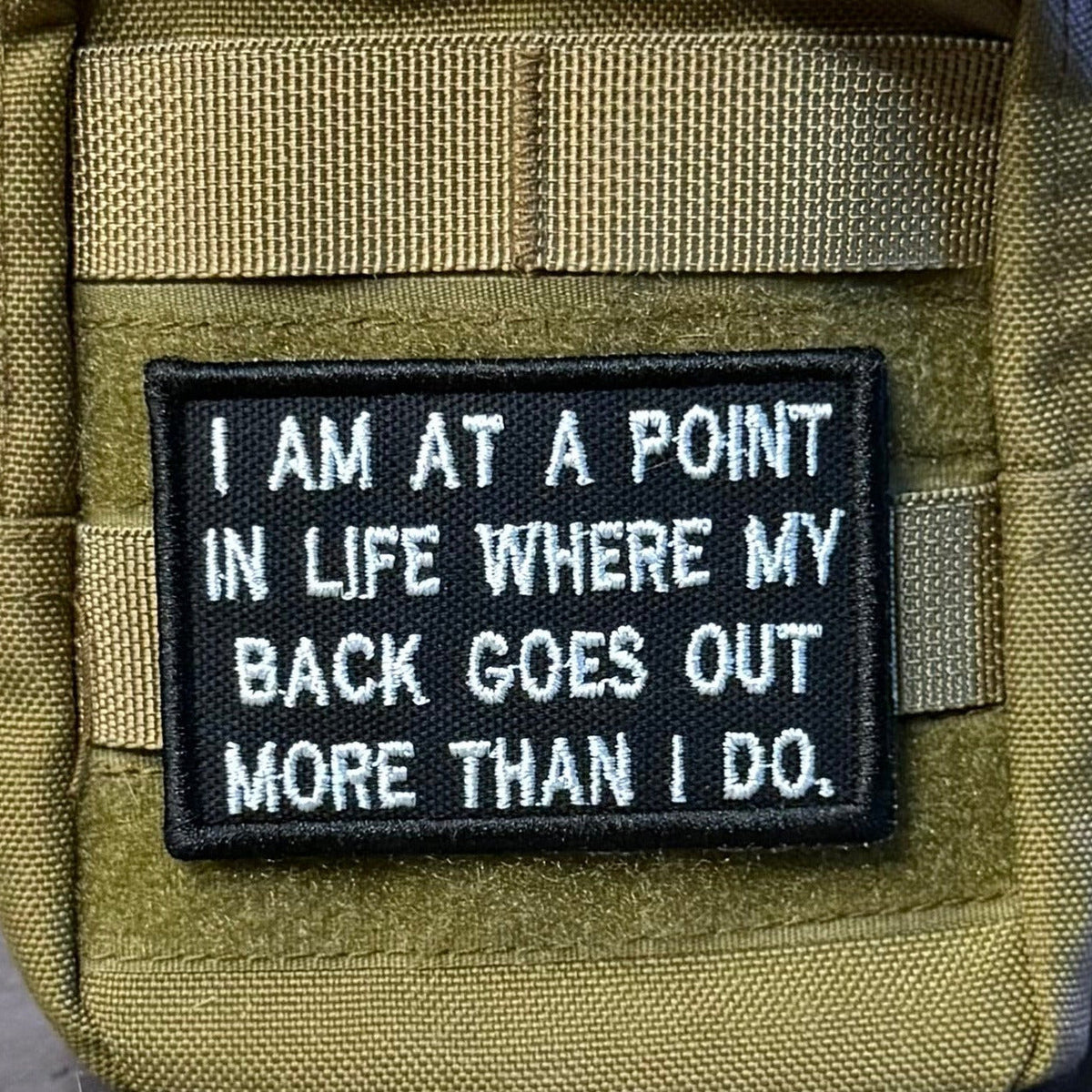 My Back Goes Out More Than I Do patch in black with white lettering, mounted on an olive backpack