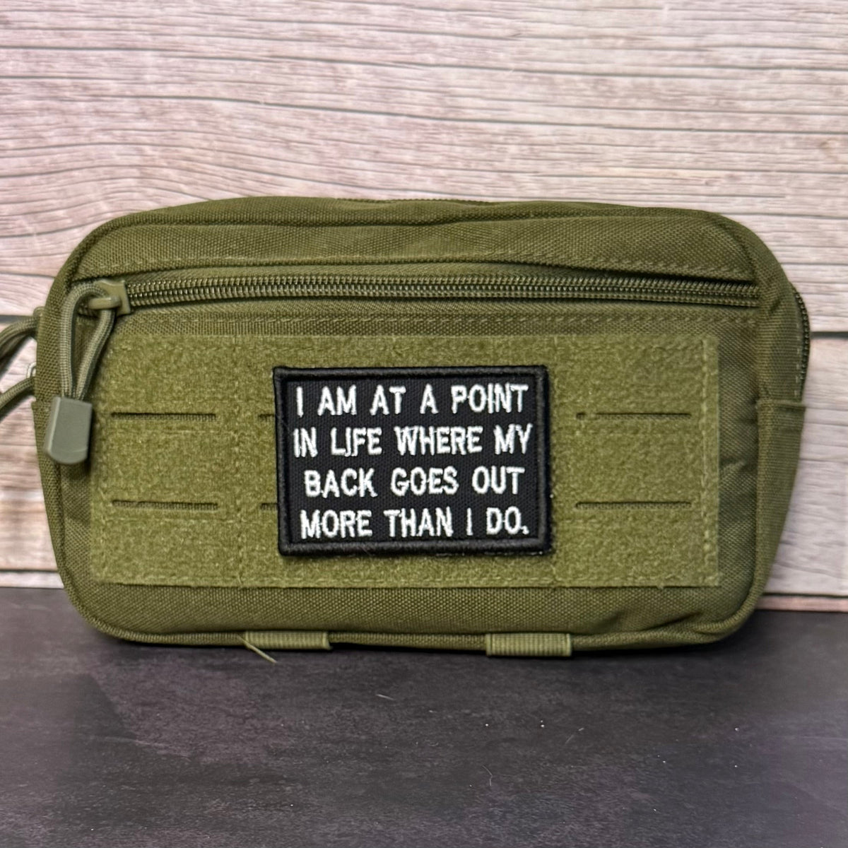 My Back Goes Out More Than I Do patch in black with white lettering, mounted on an olive utility pouch