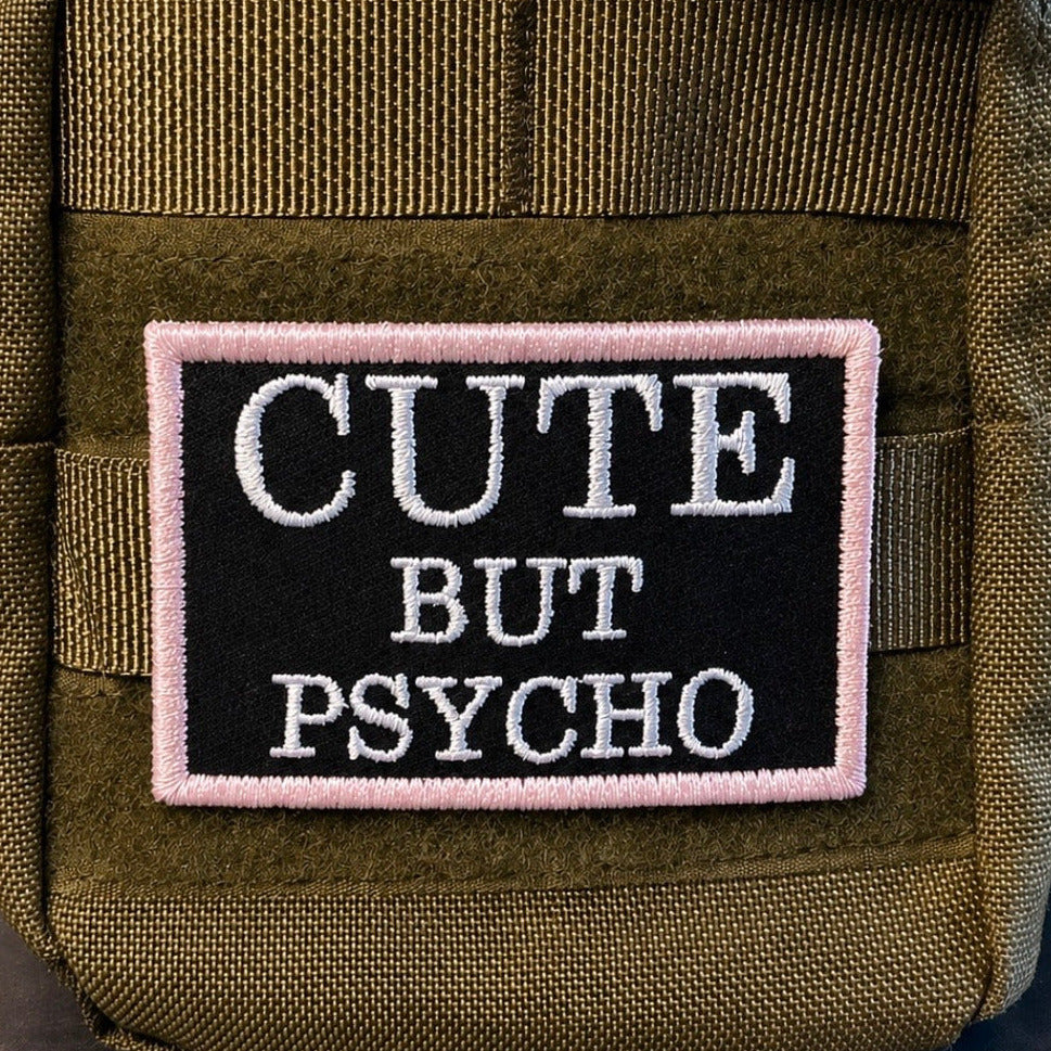 Cute But Psycho patch in black with white lettering and pale pink border, mounted on an olive backpack