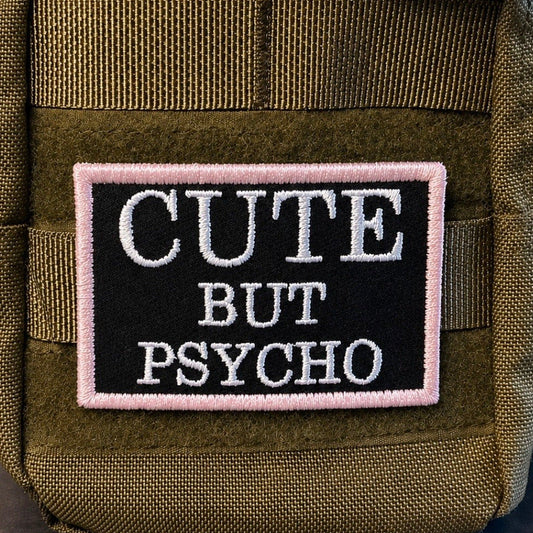 Cute But Psycho patch in black with white lettering and pale pink border, mounted on an olive backpack