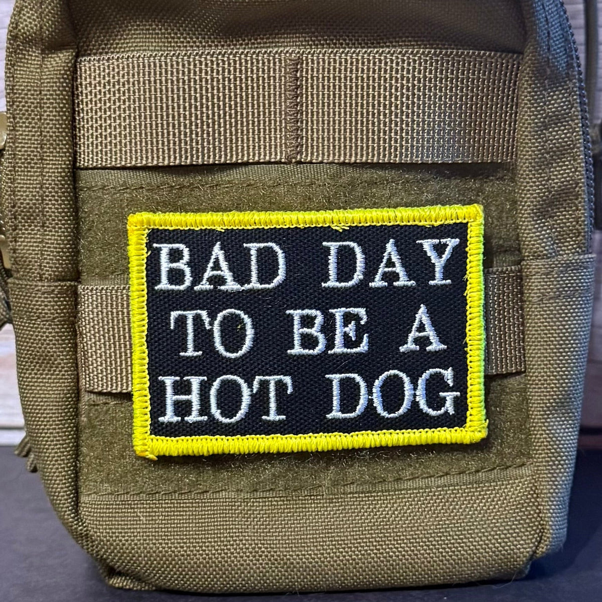 Bad Day To Be A Hot Dog patch in black with white lettering and a yellow border, mounted on an olive backpack