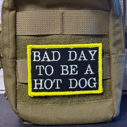 Bad Day To Be A Hot Dog patch in black with white lettering and a yellow border, mounted on an olive backpack