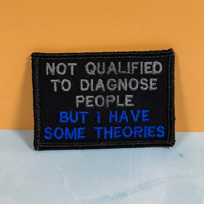 Front view of Not Qualified to Diagnose People, But I Have Theories patch in black with white and blue lettering