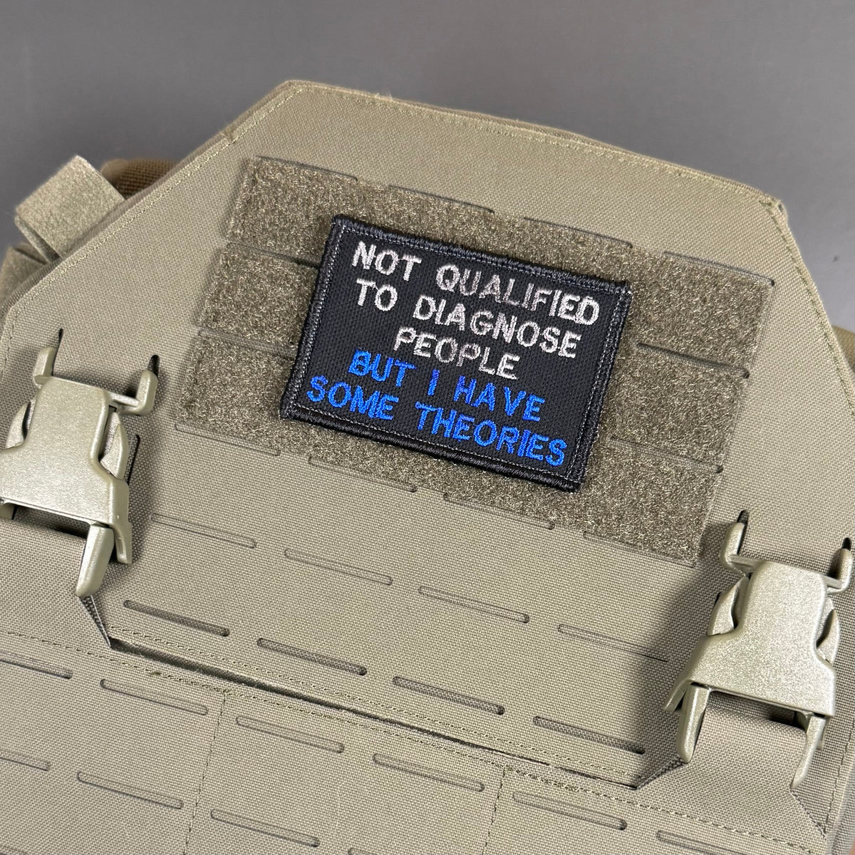 Not Qualified to Diagnose People, But I Have Theories patch in black with white and blue lettering, mounted on a tan plate carrier