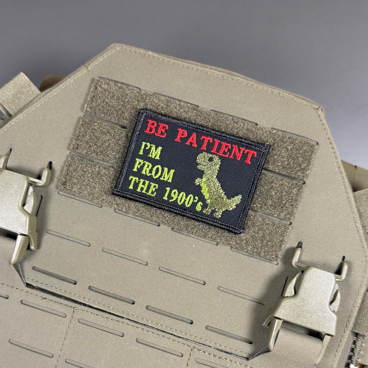 Be Patient I'm From The 1900's patch in black with red and green lettering and a green dinosaur, mounted on a tan plate carrier