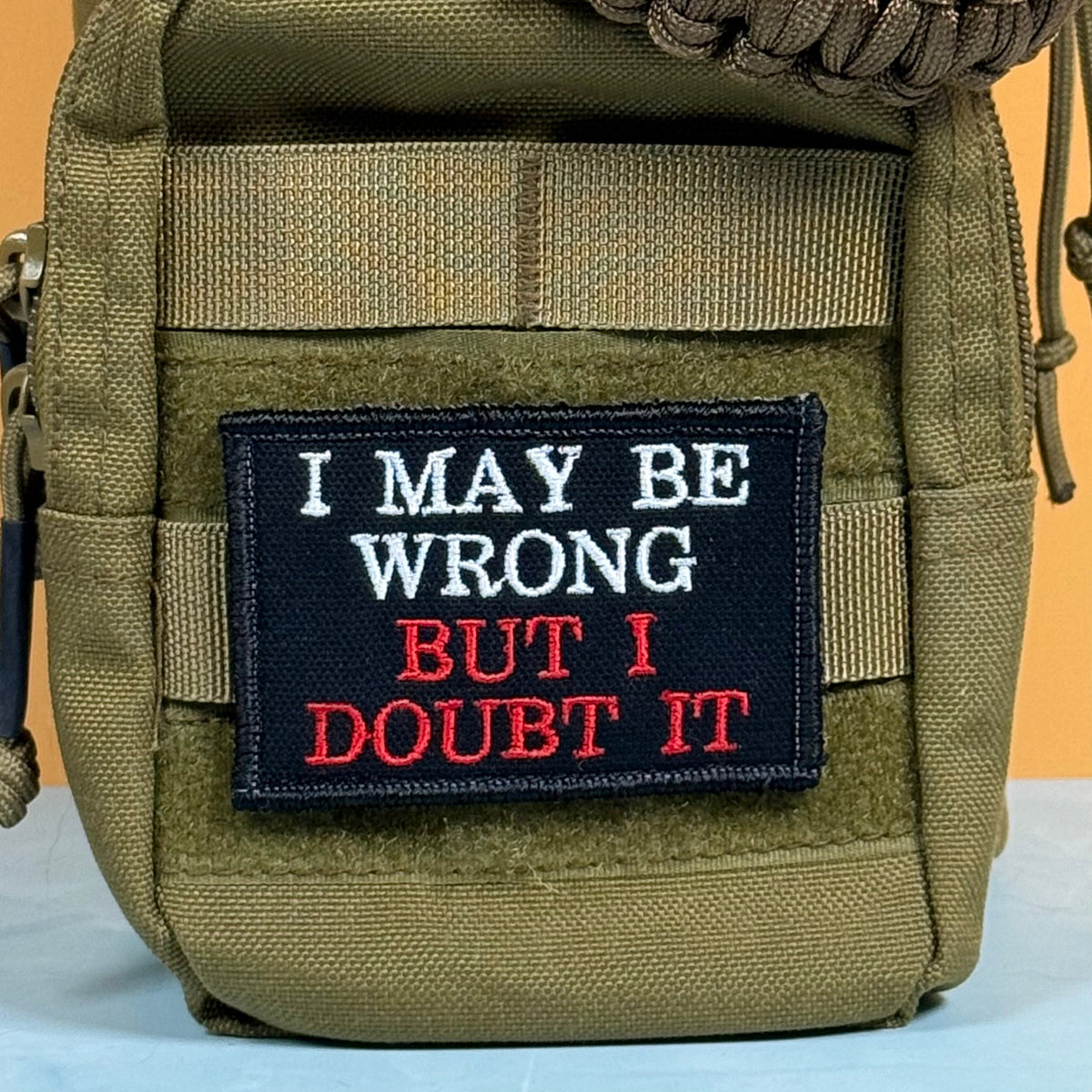 I May Be Wrong But I Doubt It patch in black with white and red lettering, mounted on an olive backpack