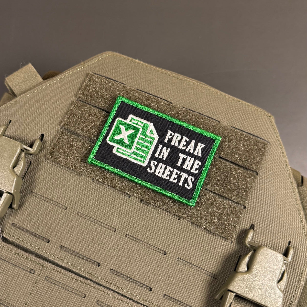 Freak In The Sheets patch in black with a green spreadsheet icon, white lettering and green border, mounted on a tan plate carrier