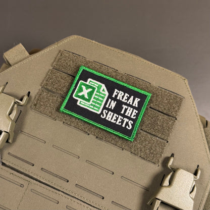 Freak In The Sheets patch in black with a green spreadsheet icon, white lettering and green border, mounted on a tan plate carrier