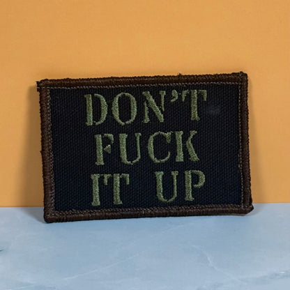 Front view of Don't Fuck It Up patch in black with olive-green lettering and a tan border