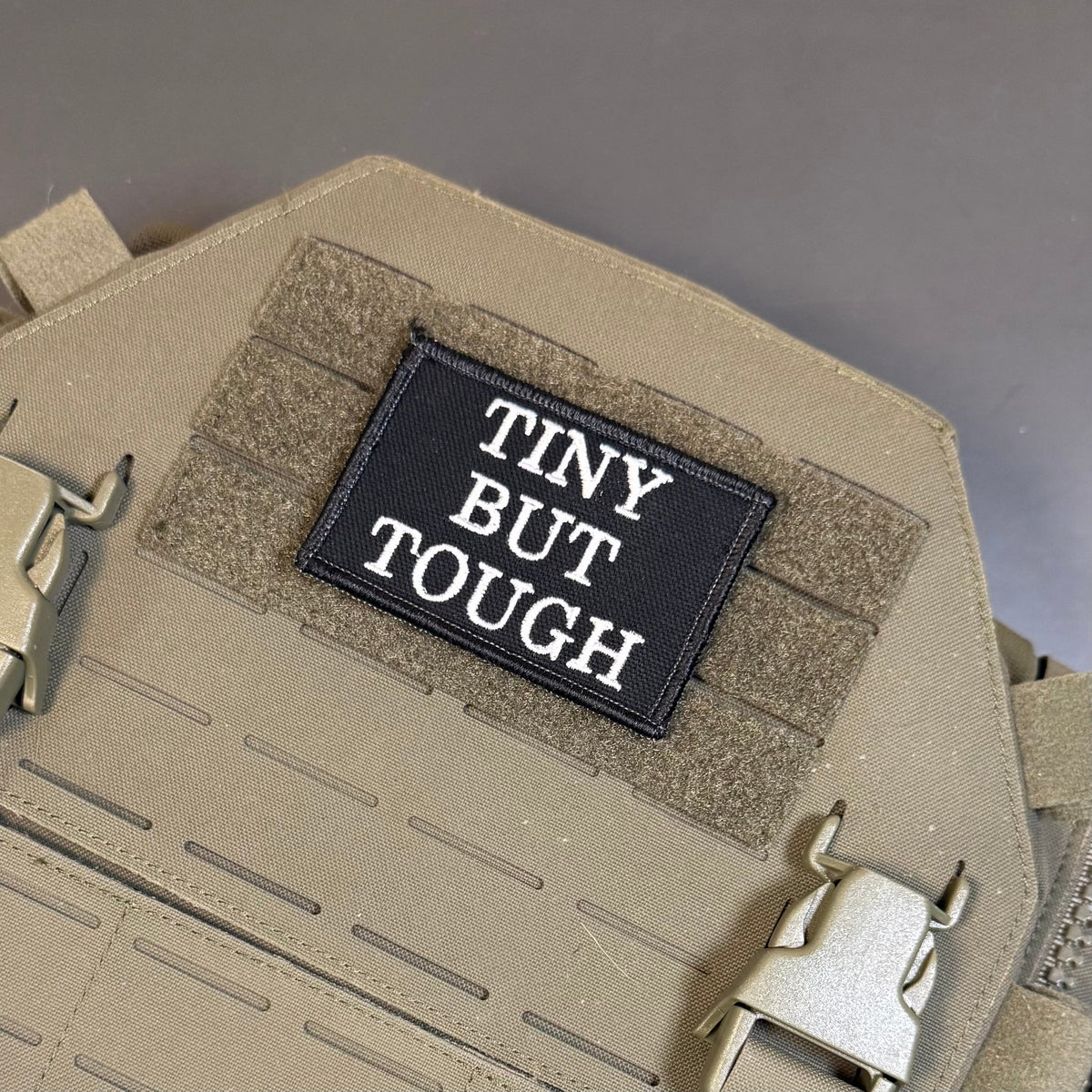 Tiny But Tough patch in black with white lettering, mounted on a tan plate carrier