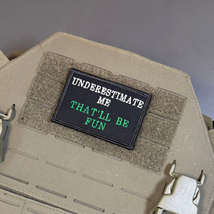 Underestimate Me, That'll Be Fun patch in black with white and green lettering, mounted on a tan plate carrier