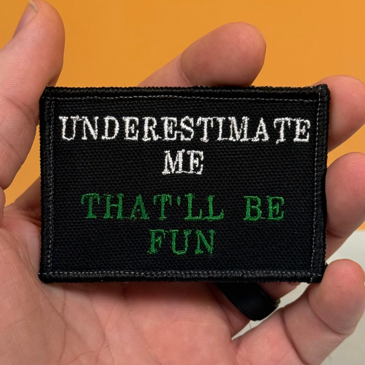 Underestimate Me, That'll Be Fun patch in black with white and green lettering, held in a hand