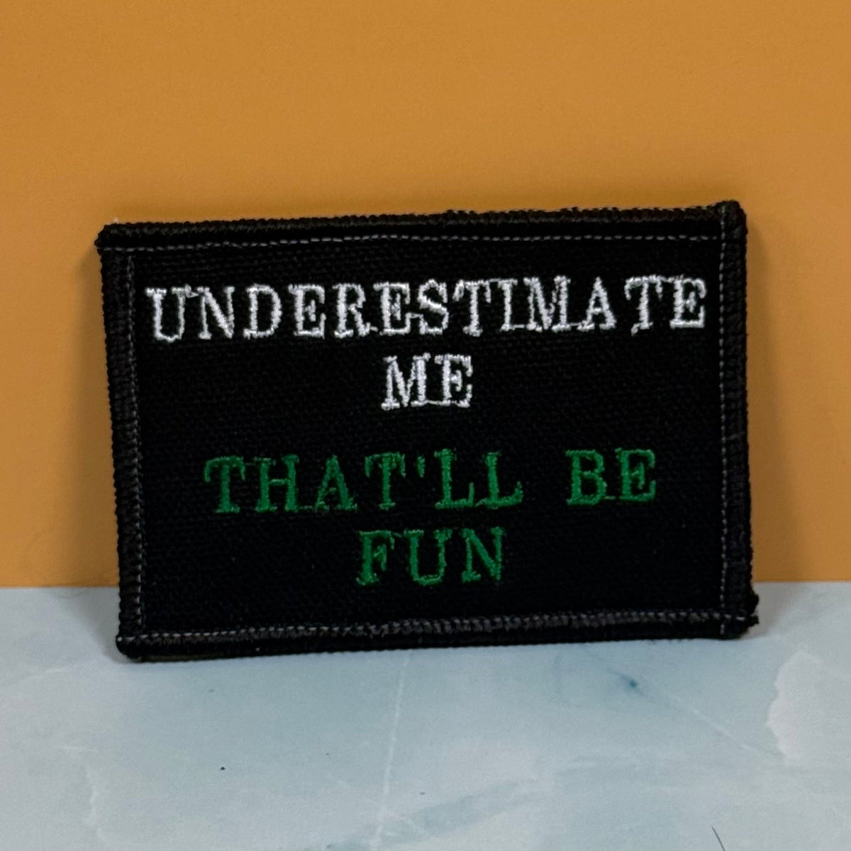 Front view of Underestimate Me, That'll Be Fun patch in black with white and green lettering