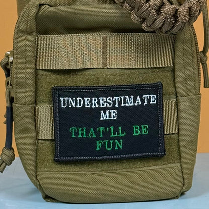 Underestimate Me, That'll Be Fun patch in black with white and green lettering, mounted on an olive backpack