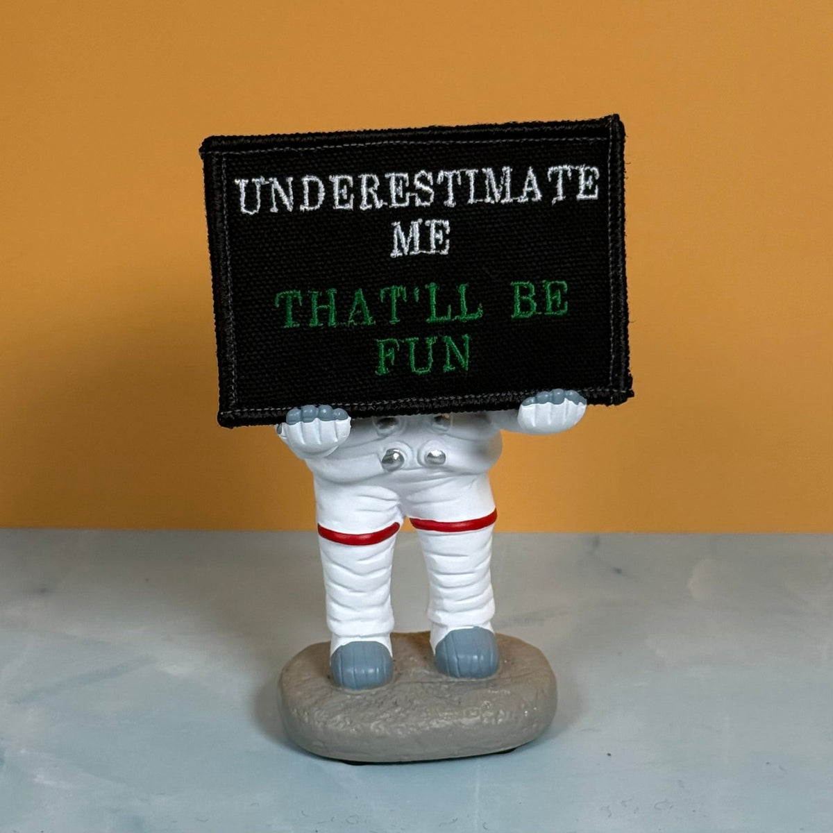 Underestimate Me, That'll Be Fun patch in black with white and green lettering, propped on a small astronaut figurine