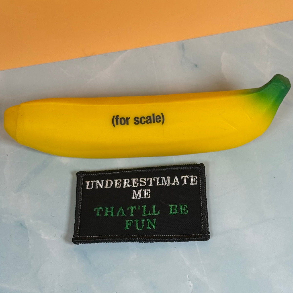 Underestimate Me, That'll Be Fun patch in black with white and green lettering, beside a yellow banana-shaped object