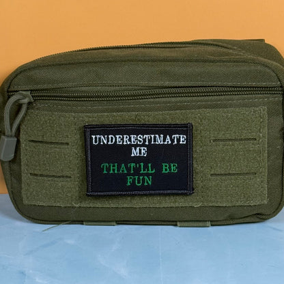 Underestimate Me, That'll Be Fun patch in black with white and green lettering, mounted on an olive utility pouch