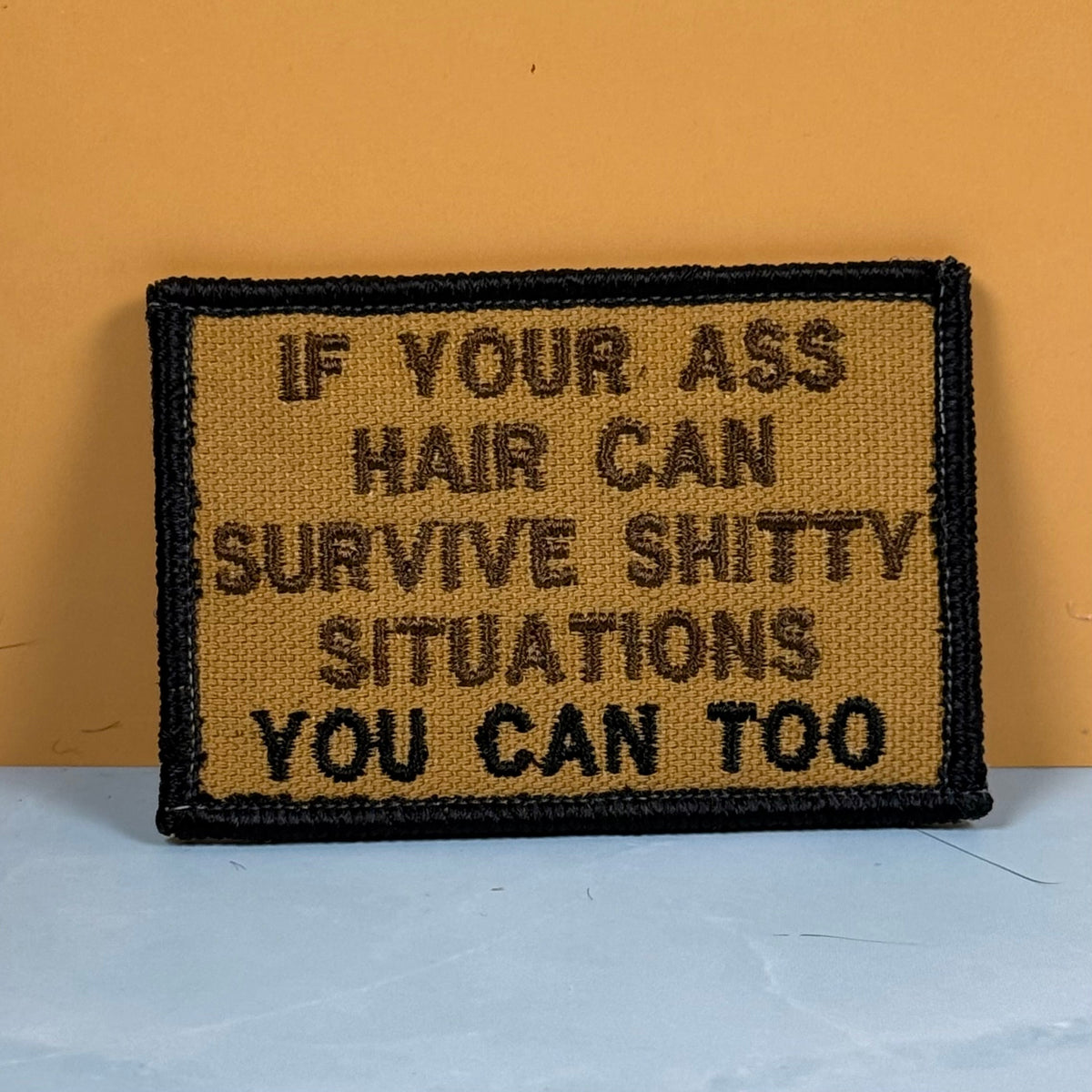 Front view of If Your Ass Hair Can Survive patch in tan with dark lettering