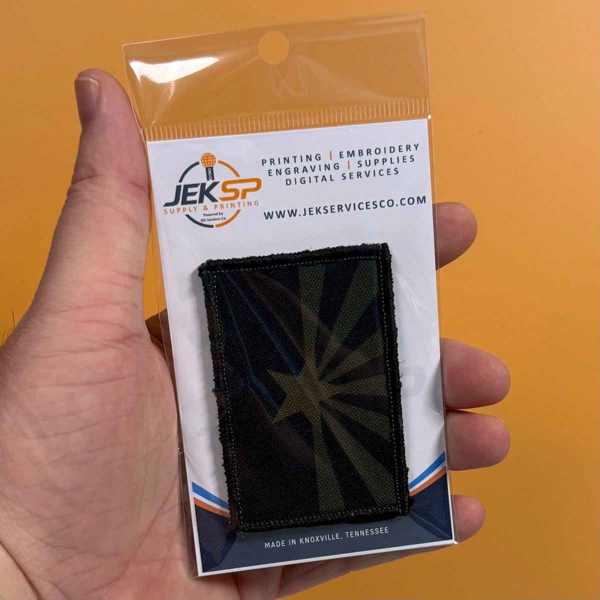 Arizona State Flag patch in subdued tan and dark blue Arizona flag with a star and radiating rays, retail package held in a hand