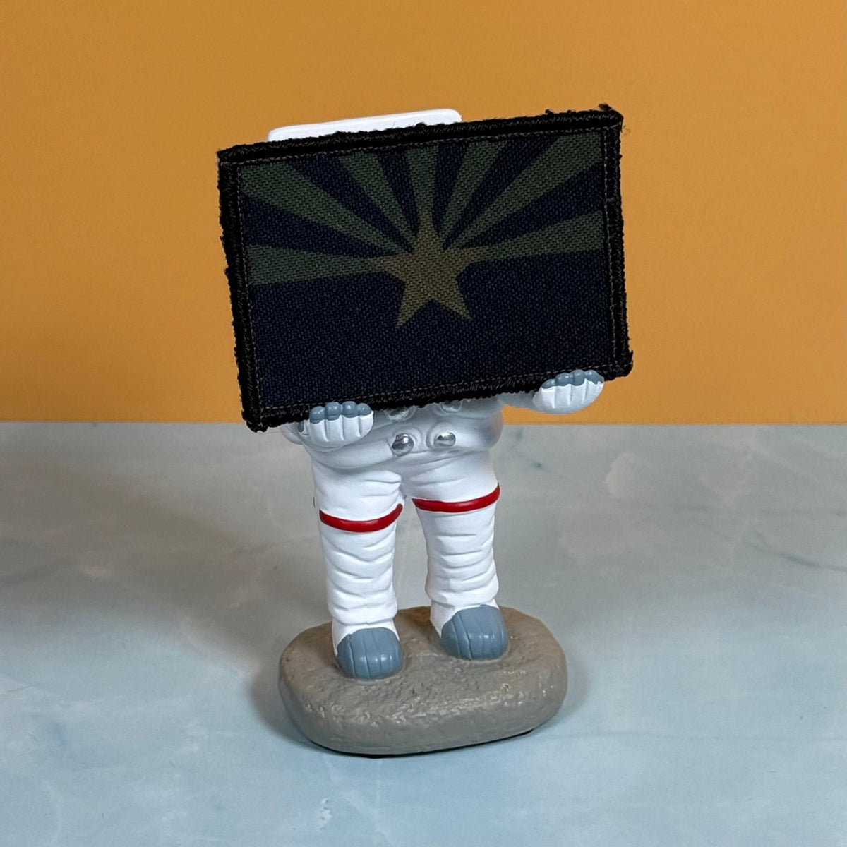 Arizona State Flag patch in subdued tan and dark blue Arizona flag with a star and radiating rays, propped on a small astronaut figurine