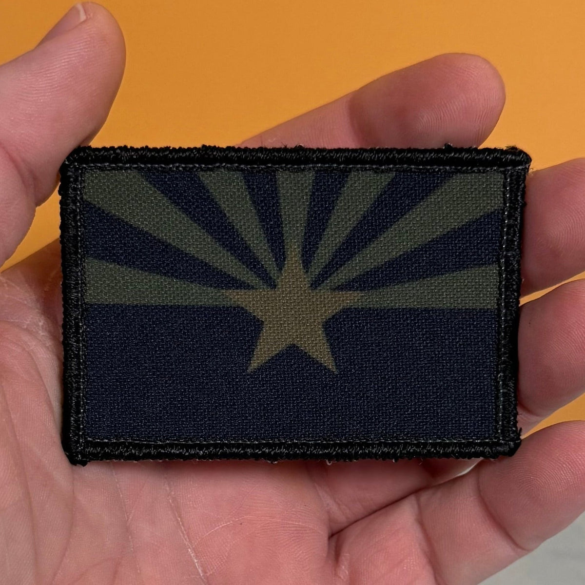 Arizona State Flag patch in subdued tan and dark blue Arizona flag with a star and radiating rays, held in a hand