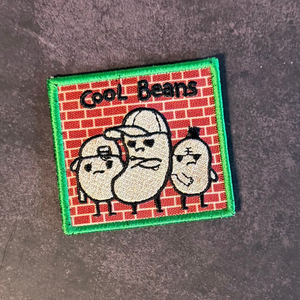 Front view of Cool Beans patch in red brick background with three cream-colored cartoon beans in sunglasses and a green border