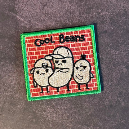 Front view of Cool Beans patch in red brick background with three cream-colored cartoon beans in sunglasses and a green border