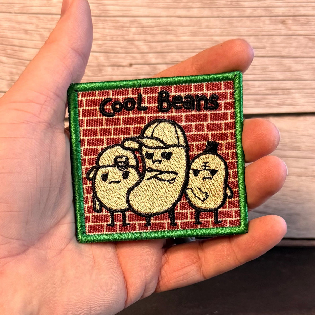 Cool Beans patch in red brick background with three cream-colored cartoon beans in sunglasses and a green border, held in a hand