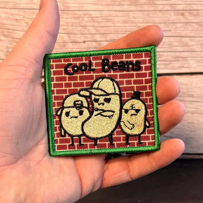 Cool Beans patch in red brick background with three cream-colored cartoon beans in sunglasses and a green border, held in a hand