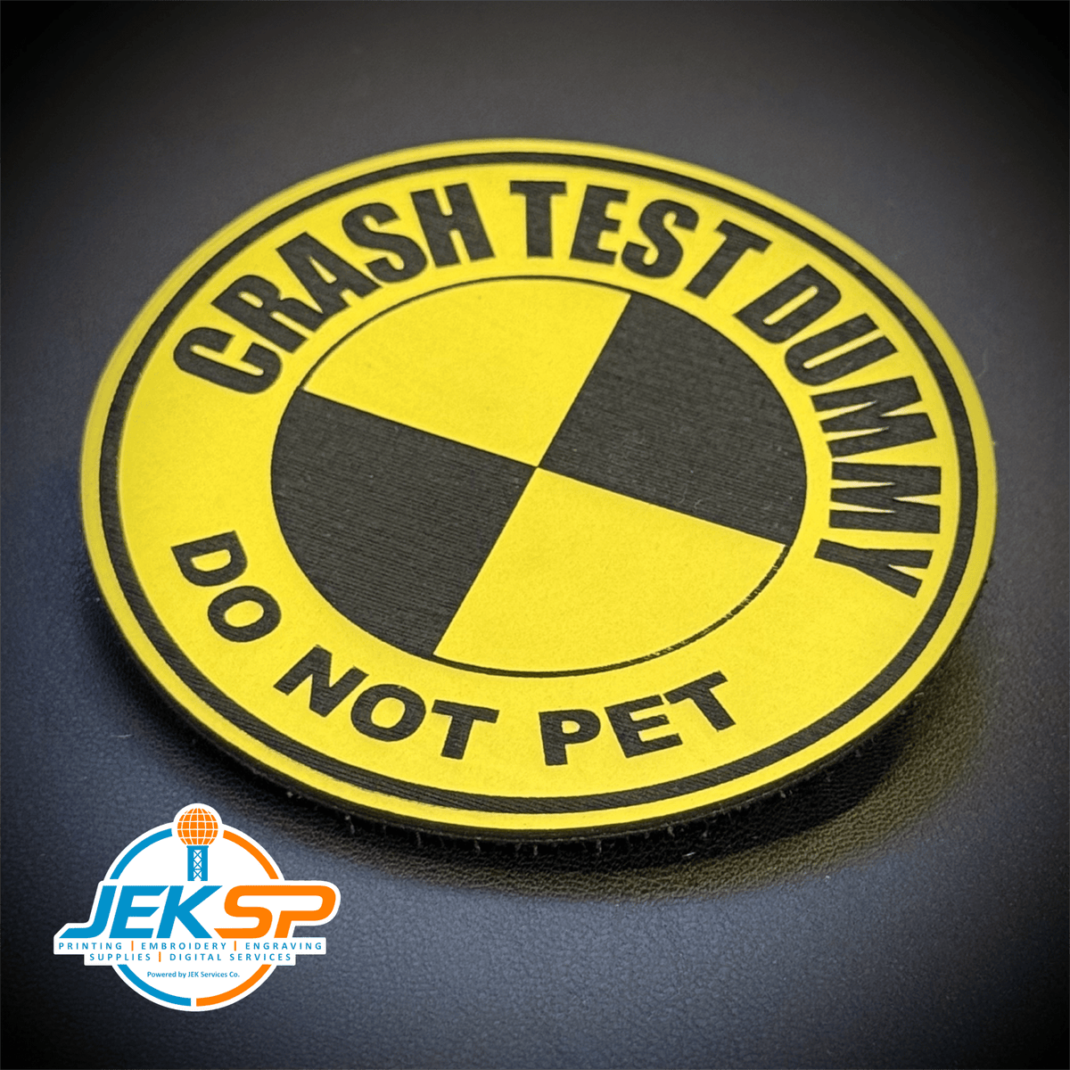 Front view of Crash Test Dummy patch in yellow with a black quartered circle and curved lettering