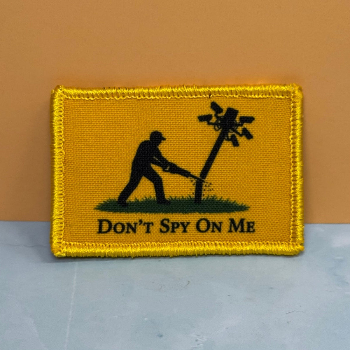 Front view of Don't Spy On Me, Anti-Flock Cameras patch in yellow with a black figure kicking a camera pole and black lettering