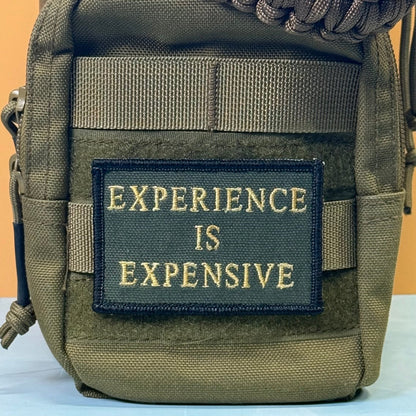 Experience Is Expensive patch in olive green with yellow lettering, mounted on an olive backpack