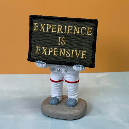 Experience Is Expensive patch in olive green with yellow lettering, propped on a small astronaut figurine